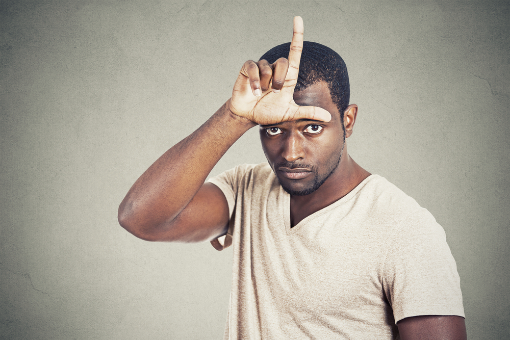 Closeup portrait serious young man showing loser sign on forehead looking at you with disgust at camera isolated grey wall background. Negative human emotion facial expression feeling